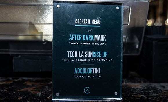A printed After Dark cocktail menu in an acrylic stand on the bar, listing drinks named for the campaign.