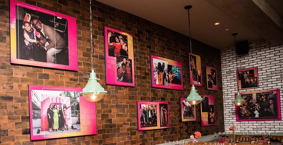 The Still Rising gallery wall: framed event photographs in pink and yellow mounts running along an exposed brick wall.